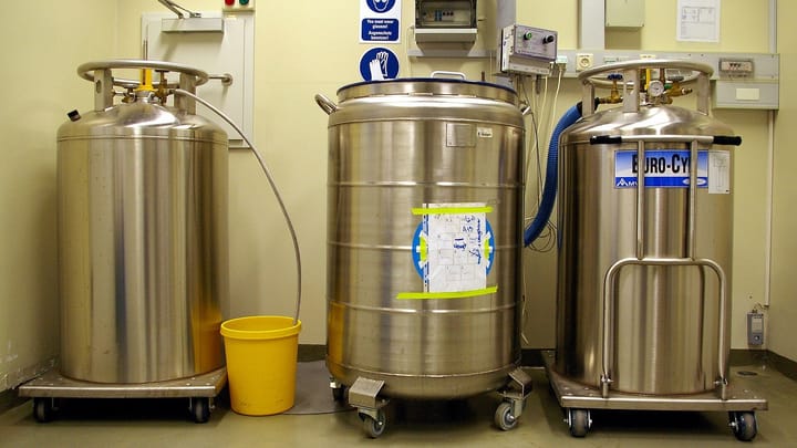 Three stainless steel liquid nitrogen tanks of the kind used for frozen biological samples, standing on wheeled bases in a laboratory storage room