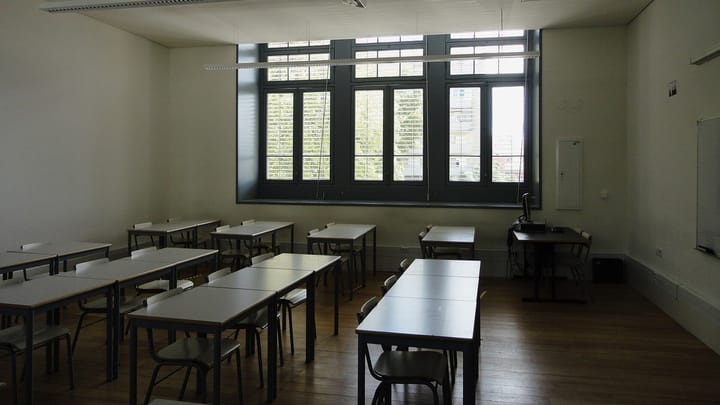 An empty secondary school classroom in Lisbon with rows of white desks and chairs on a wooden floor, facing tall shuttered windows