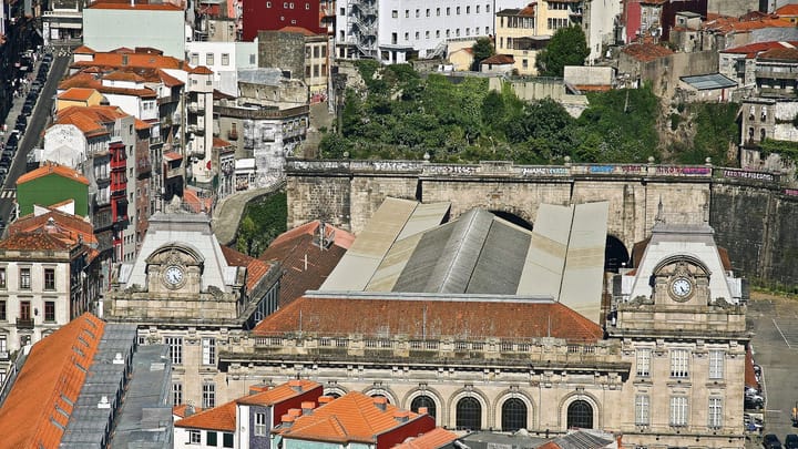 São Bento railway station in Porto seen from above, its granite facade and clock towers among orange tiled roofs, with the train shed and tunnel mouths behind